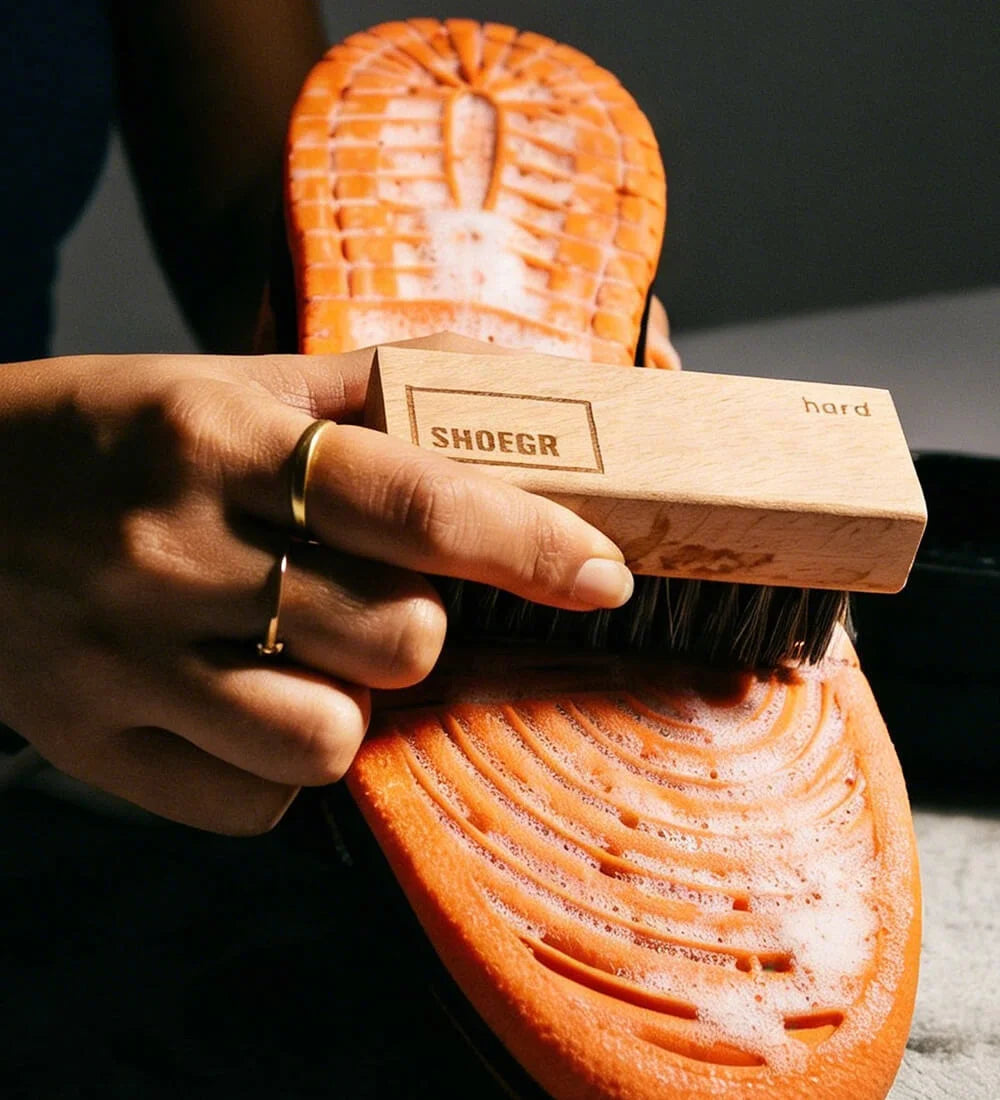 Person cleaning an orange sneaker outsolewith the SHOEGR Hard Shoe Cleaning Brush